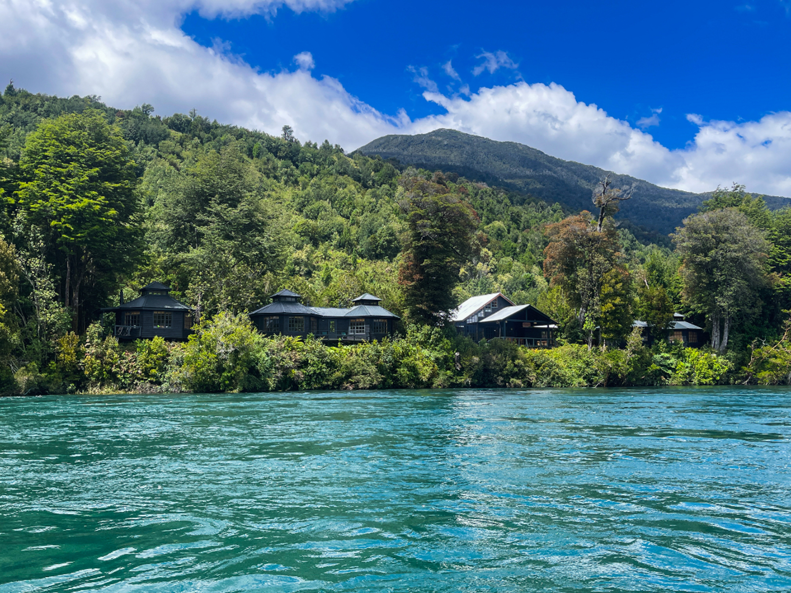 A lodge sits on a river bank with lush trees and greenery surrounding the lodge and bright blue rive