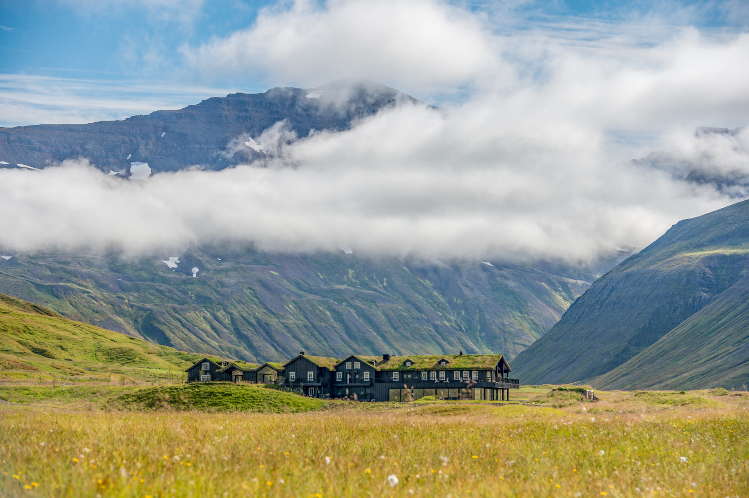 A large lodge is seen with clouds above, mountains behind on a summer day. The lodge has a dark exte