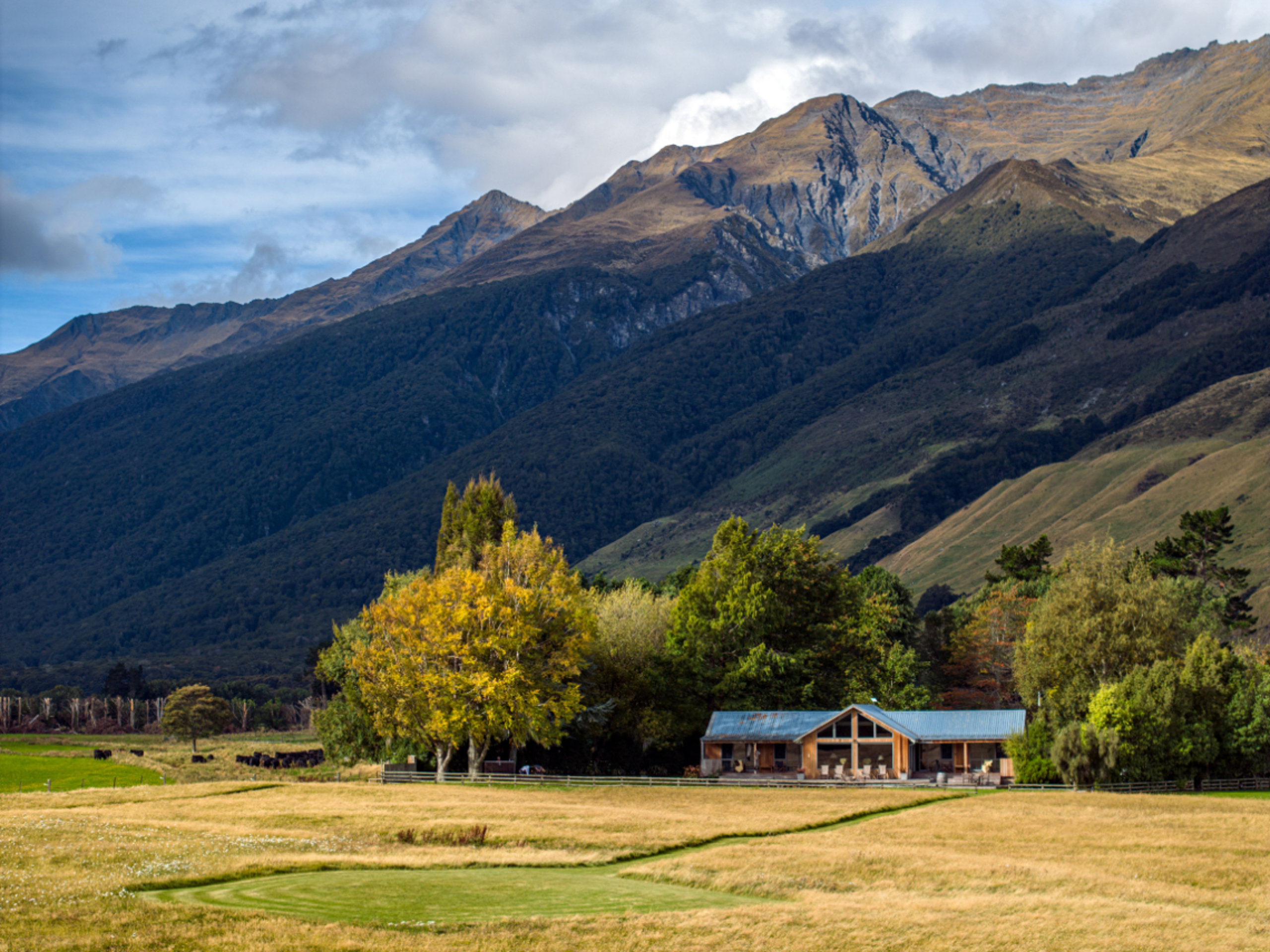 A lodge sits in the center of a field with mountains behind it. Trees surround the lodge on a sunny 