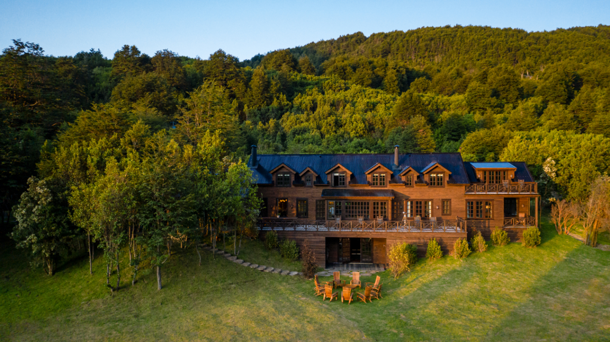 A warm colored wood lodge sits at the base of a mountain, with a large lawn and chairs in front on a