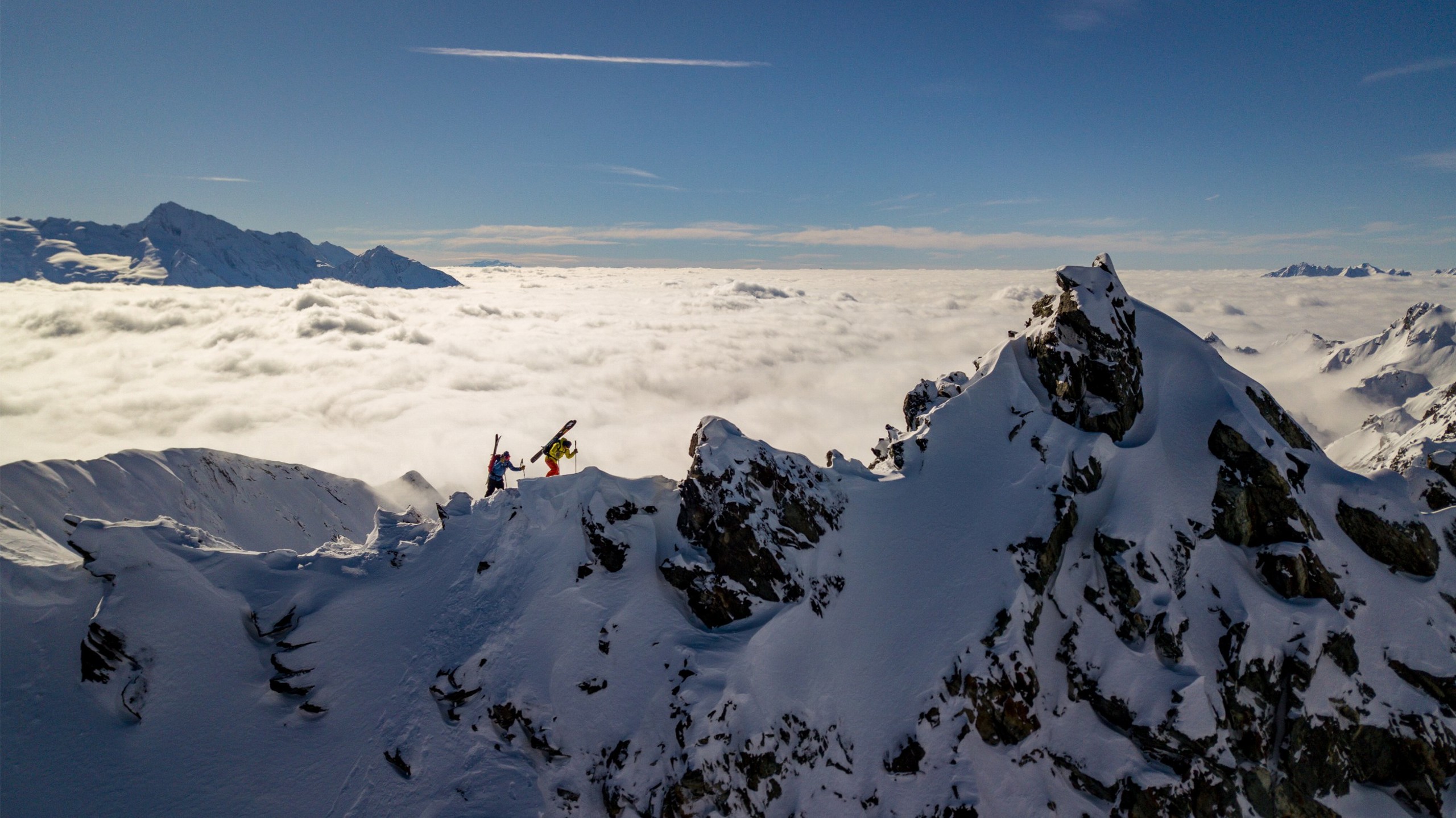 Two skiers walking across the ridge of a mountain above the clouds.
