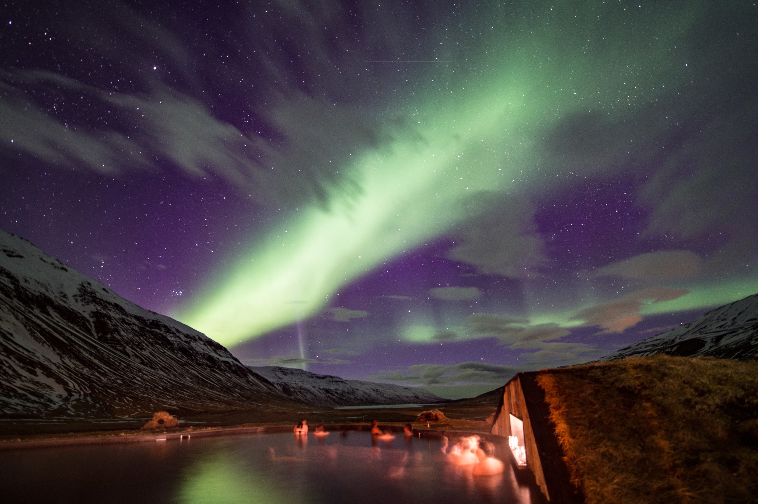 A group of people in an outdoor heated pool under the northern lights in the night sky.
